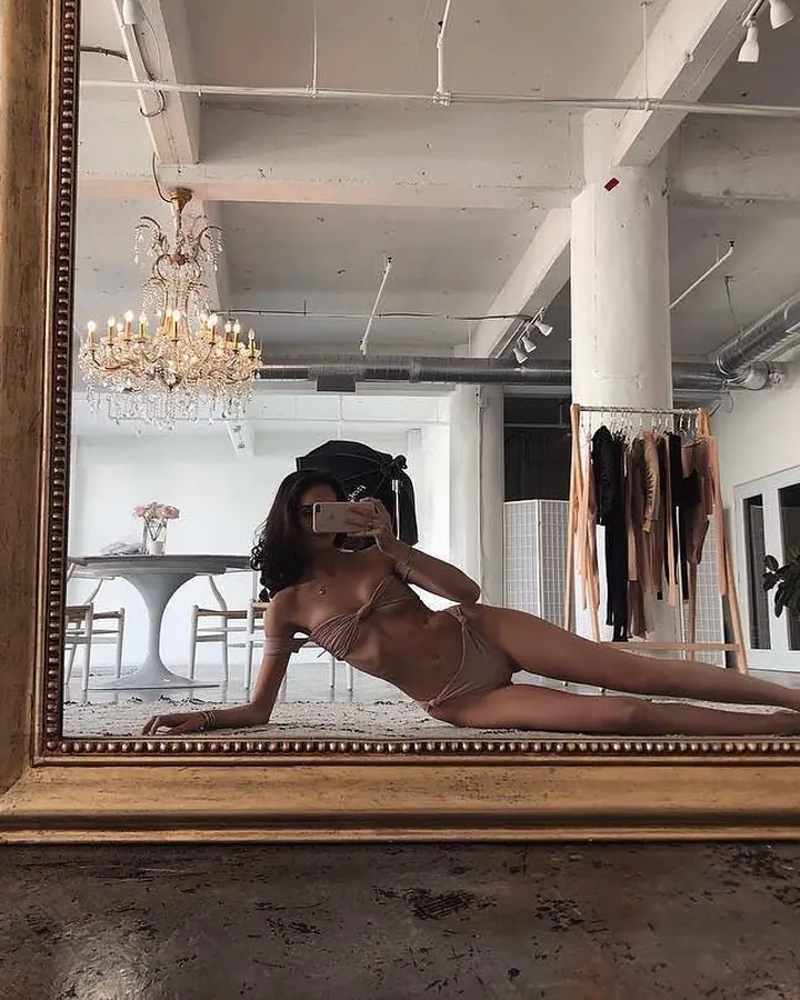 Femme en maillot de bain beige prenant un selfie dans un grand miroir doré dans un loft lumineux.