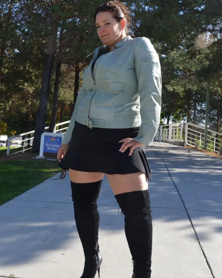 Femme en veste grise et jupe noire avec bottes hautes posant sur un trottoir ensoleillé.
