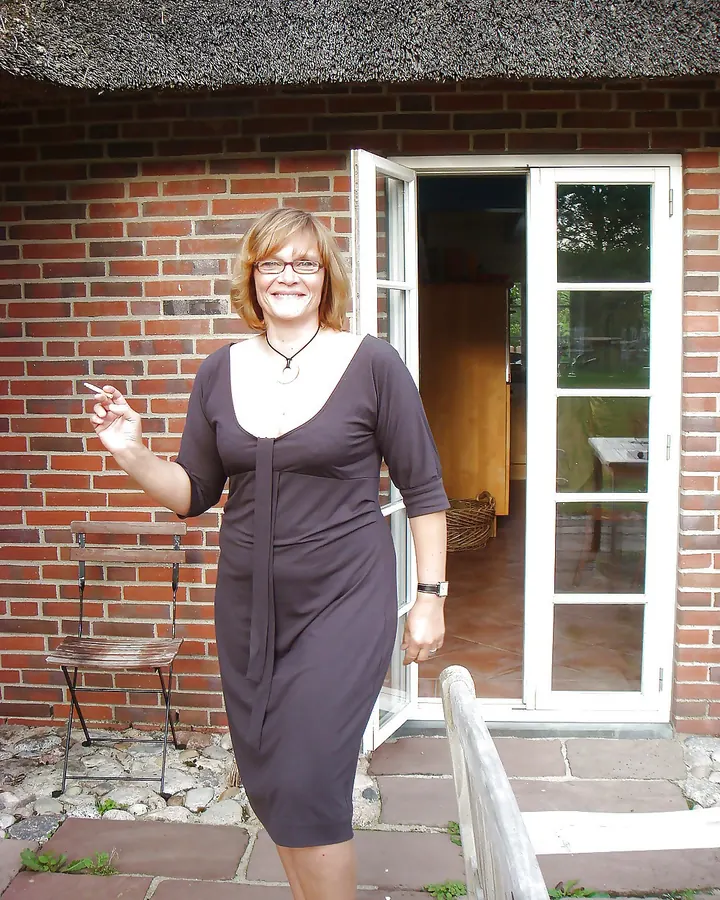 Femme aux cheveux blonds en robe marron, tenant une cigarette, devant une maison en briques avec toit de chaume.