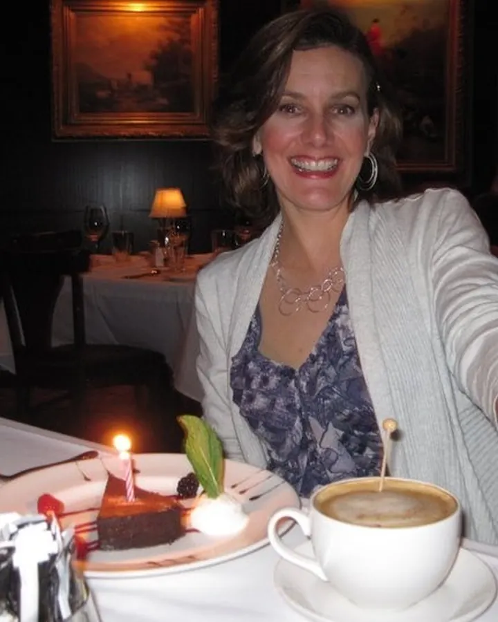 Femme souriante en tenue de soirée, avec un gâteau d'anniversaire et une tasse de café sur une table.
