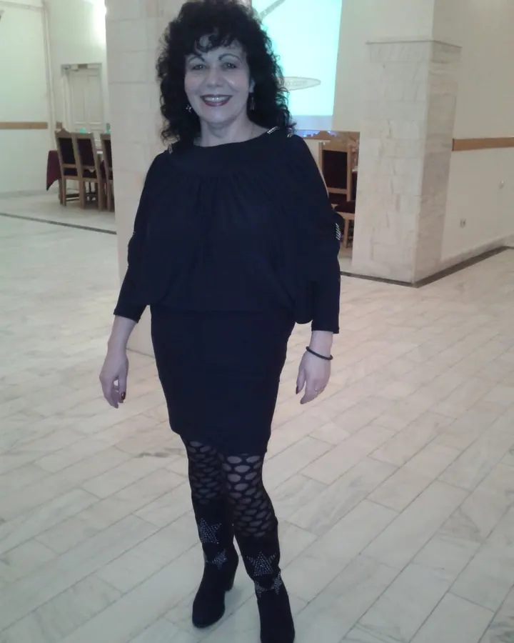 Femme européenne aux cheveux bruns-noirs, portant un haut noir et un string noir, assise sur un canapé en cuir, regardant vers l'appareil.