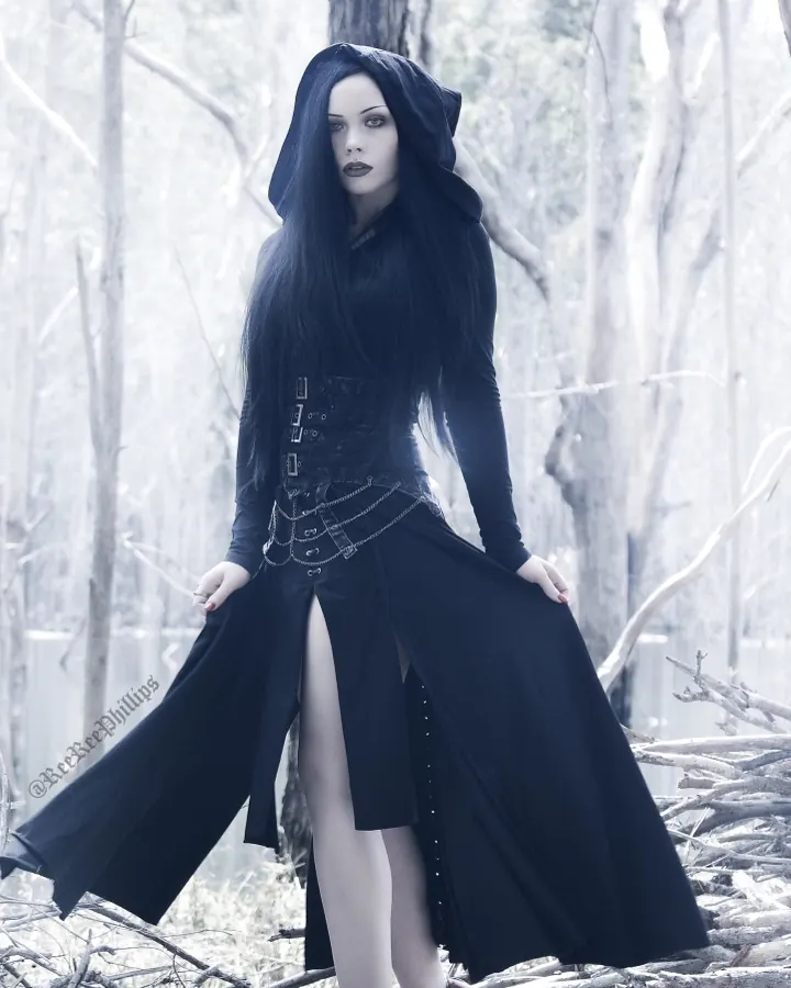 Femme aux cheveux noirs portant une robe noire à capuche, debout dans une forêt morte
