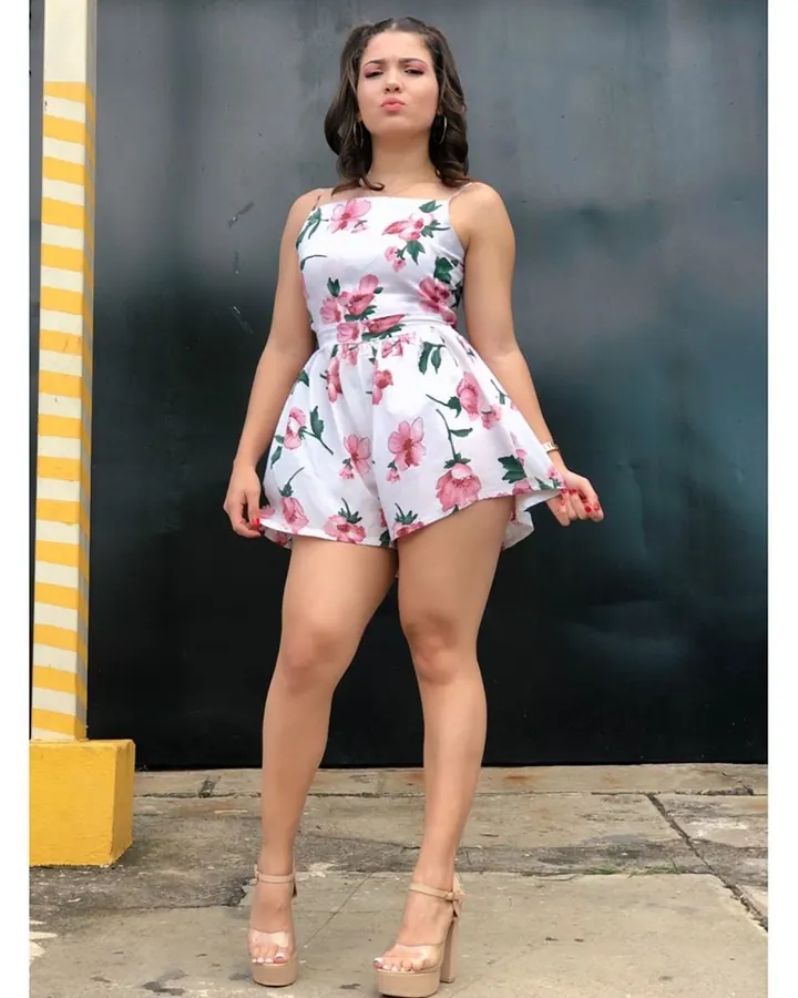 Jeune femme brune en robe fleurie blanche, posant devant un mur sombre, pieds nus en sandales à talons.