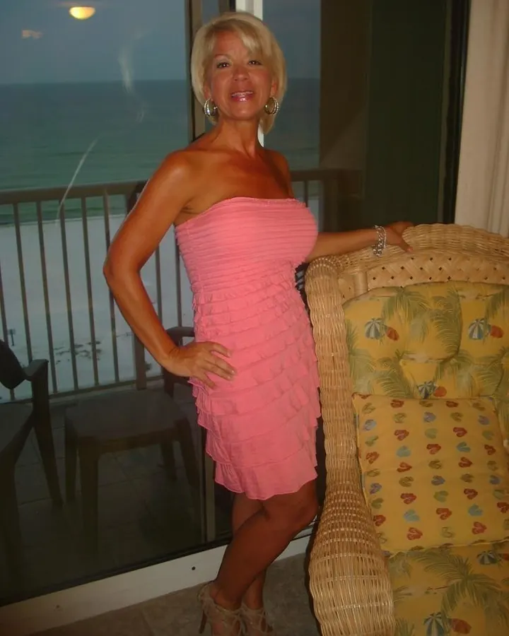 Femme blonde aux cheveux courts, robe rose, souriante, posant près d’un canapé devant une baie vitrée donnant sur la mer.