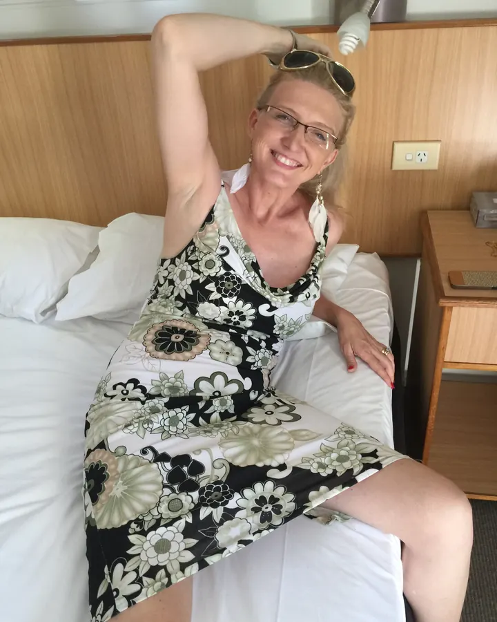Femme blonde aux cheveux courts, robe fleurie, souriante, assise sur un lit dans une chambre d’hôtel.
