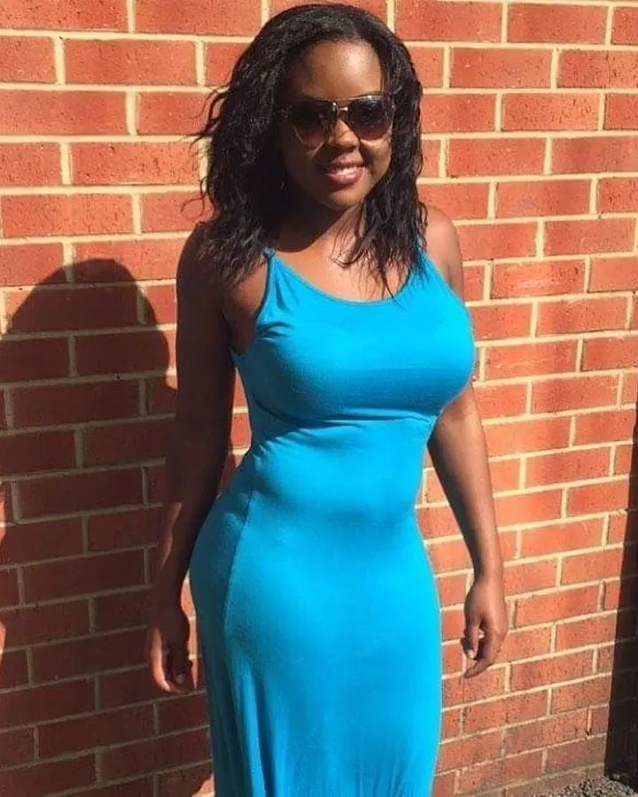 Femme africaine aux cheveux bruns-noirs, debout contre un mur en briques, portant une robe bleue, avec des lunettes de soleil.