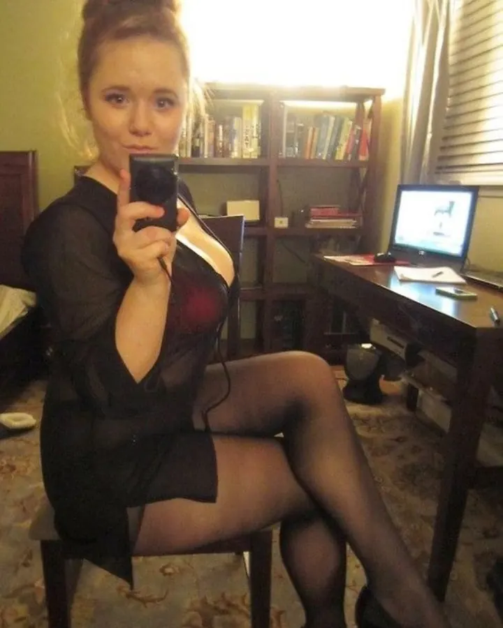 Jeune femme blonde en robe noire transparente, assise sur une chaise, tenant un téléphone portable, dans une chambre avec étagère de livres et ordinateur.