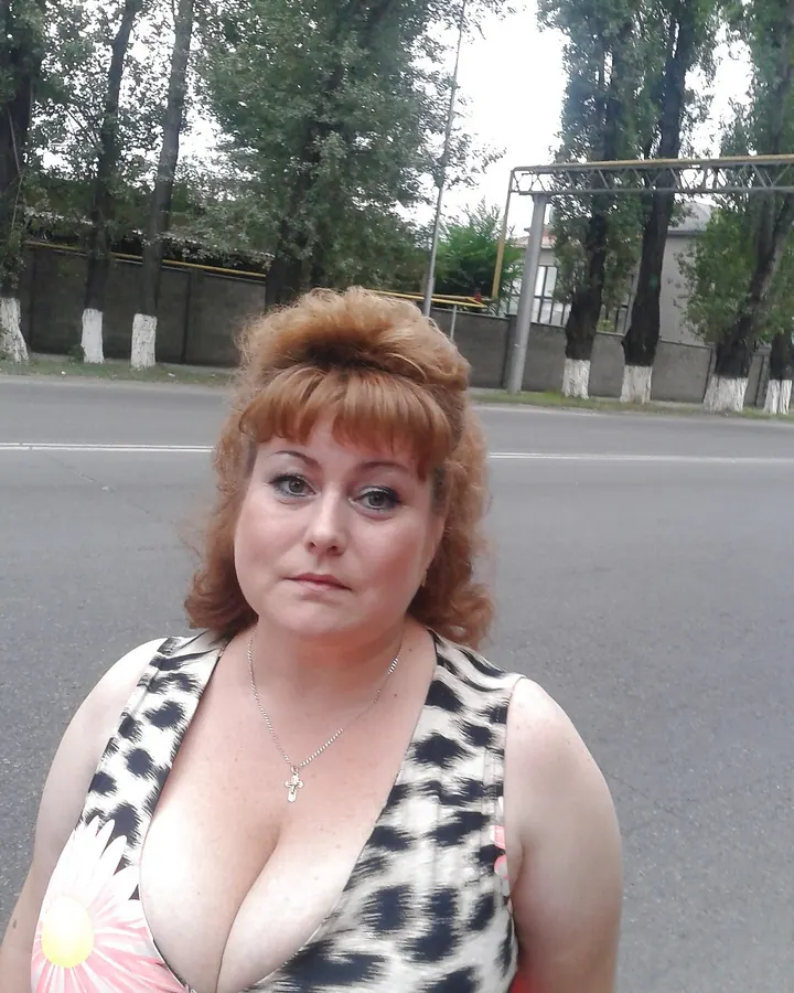 Femme mûre aux cheveux châtains, assise sur un canapé, portant un haut blanc et un short blanc, avec un collier de perles.