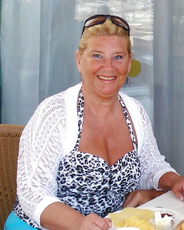 Femme mûre aux cheveux blonds, en haut blanc, posant sur une terrasse.