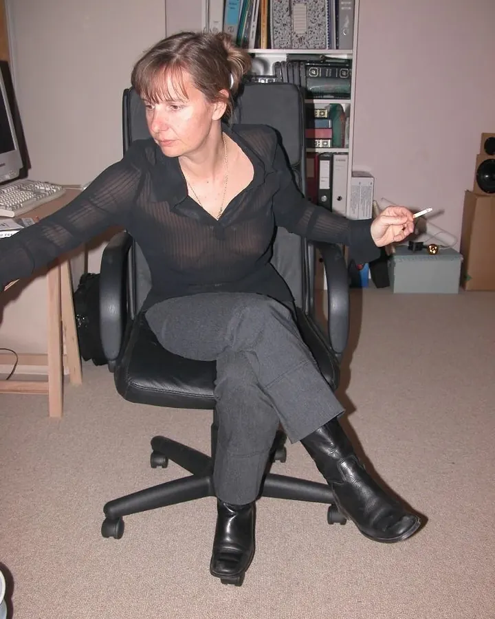 Femme assise dans un fauteuil de bureau, portant une chemise noire transparente et un pantalon gris, tenant une cigarette.