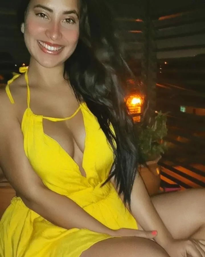Femme aux cheveux bruns portant un maillot de bain jaune, souriant, assise, cheveux longs, fond sombre.