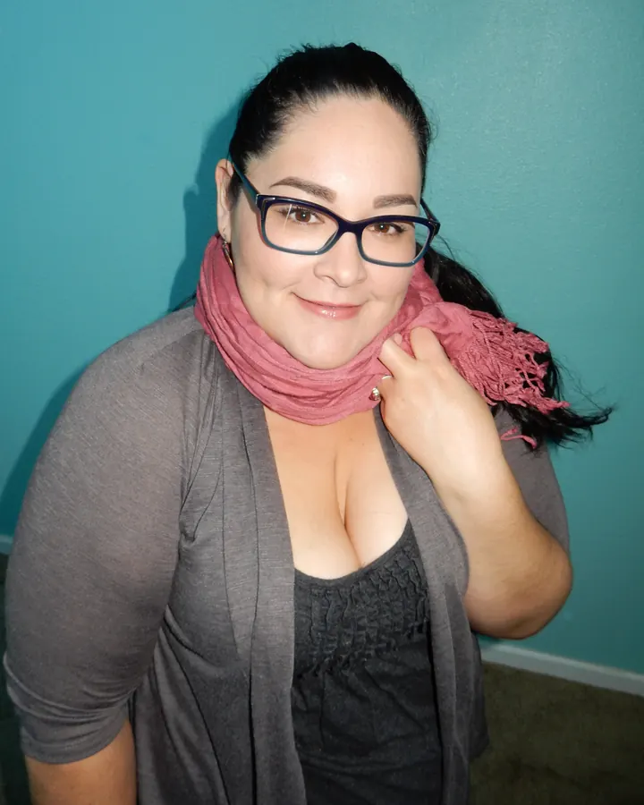 Femme aux cheveux noirs portant un haut gris, une écharpe rose, et des lunettes, souriant devant un mur bleu.