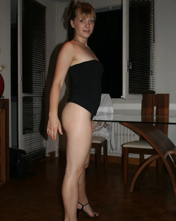 Femme européenne en haut noir sans bretelles, pose dans une pièce avec table en verre et chaises en bois.