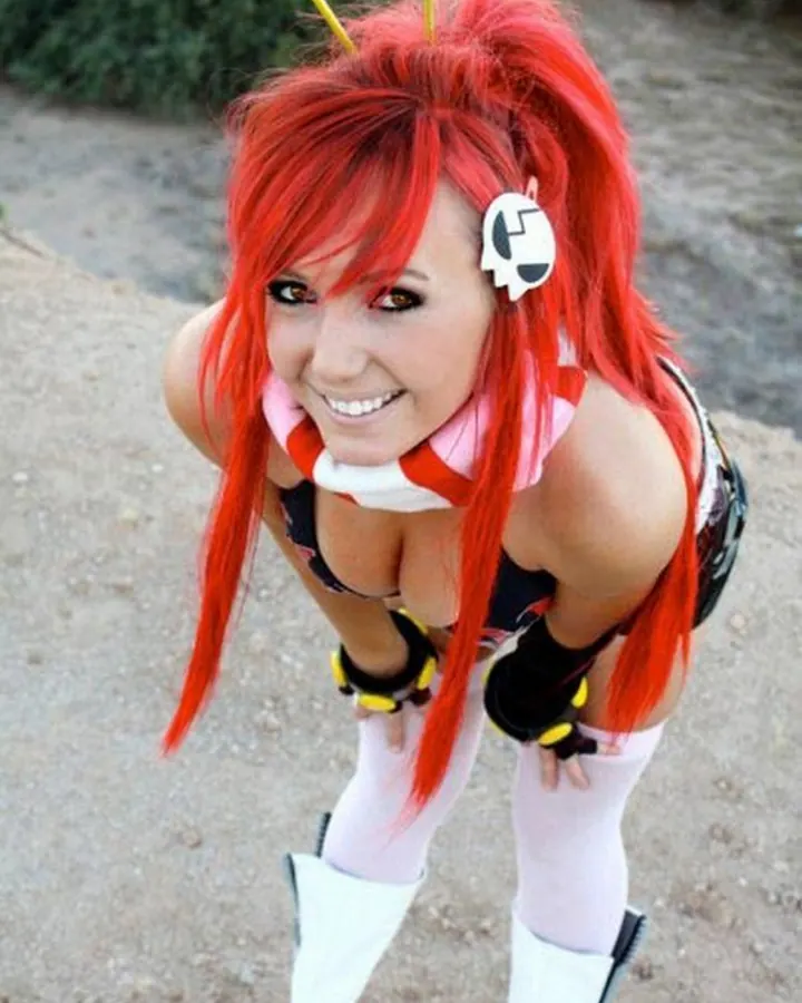 Femme aux cheveux roux, souriante, portant un costume de cosplay rouge et noir, avec des oreilles blanches et des gants noirs et jaunes.