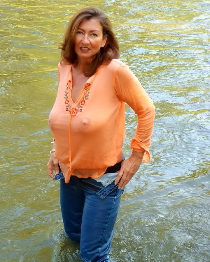 Femme blonde aux cheveux courts, souriante, portant une robe orange transparente, debout dans l'eau.