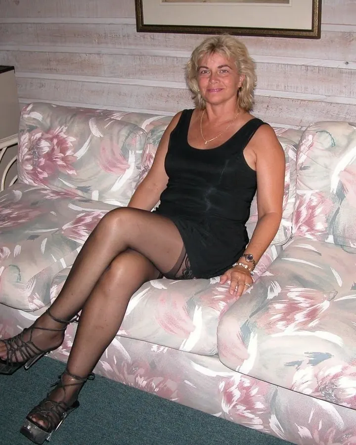 Femme aux cheveux blancs courts, en haut noir, assise sur un canapé floral, chaussée de sandales noires.