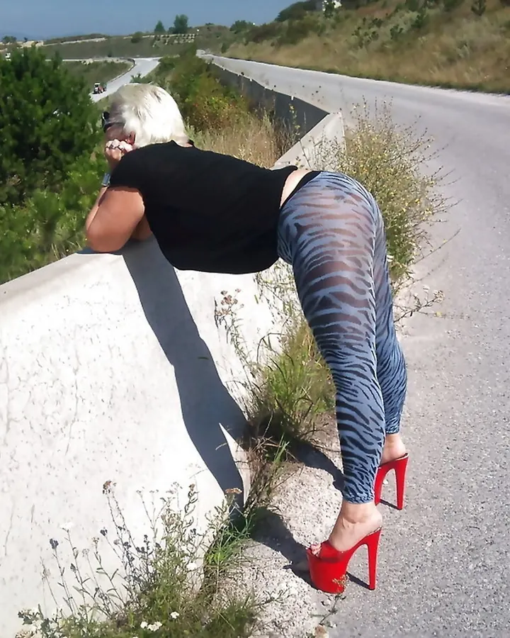 Femme blonde en collant zèbre et talons rouges, penchée sur un mur en bordure de route.