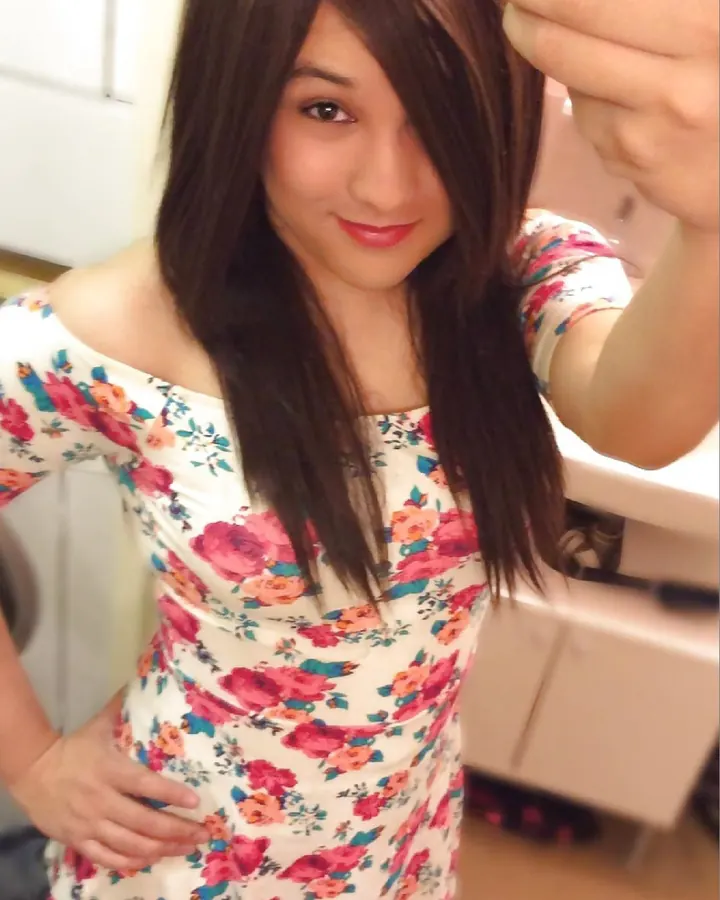 Jeune femme latine aux cheveux châtains, souriante, prenant un selfie dans un miroir, vêtue d'une robe fleurie blanche avec des roses rouges et bleues.