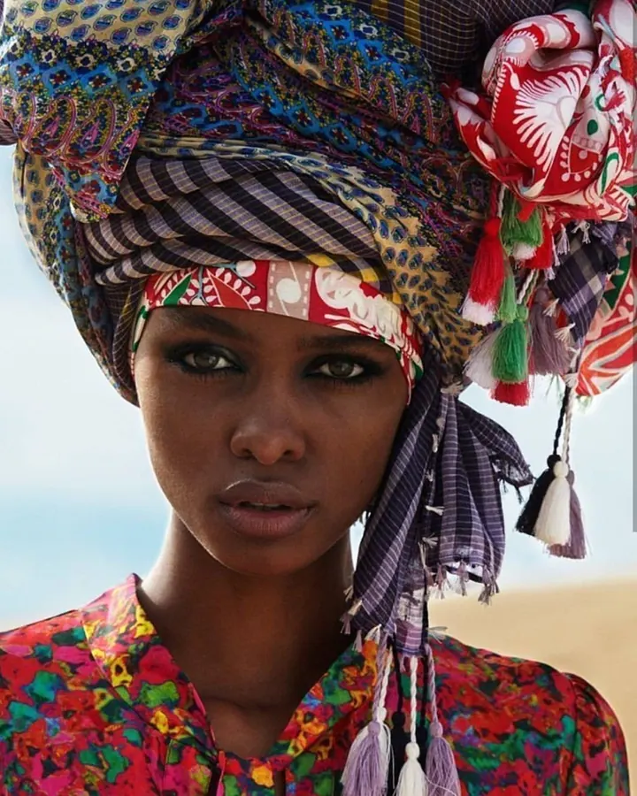 Femme africaine aux cheveux noirs, portant un turban coloré et une robe multicolore, posant en extérieur avec un ciel bleu en arrière-plan.