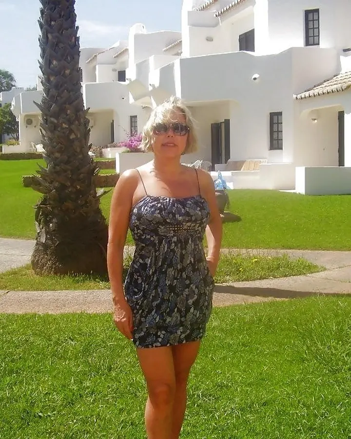 Femme aux cheveux blonds courts en robe imprimée bleu foncé, lunettes de soleil, debout sur une pelouse verte devant des bâtiments blancs.