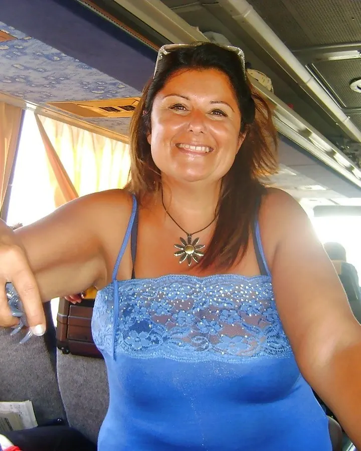 Femme aux cheveux châtains en haut noir, jupe en jean courte, veste en cuir noir, chaussures noires, appuyée sur une voiture argentée la nuit.