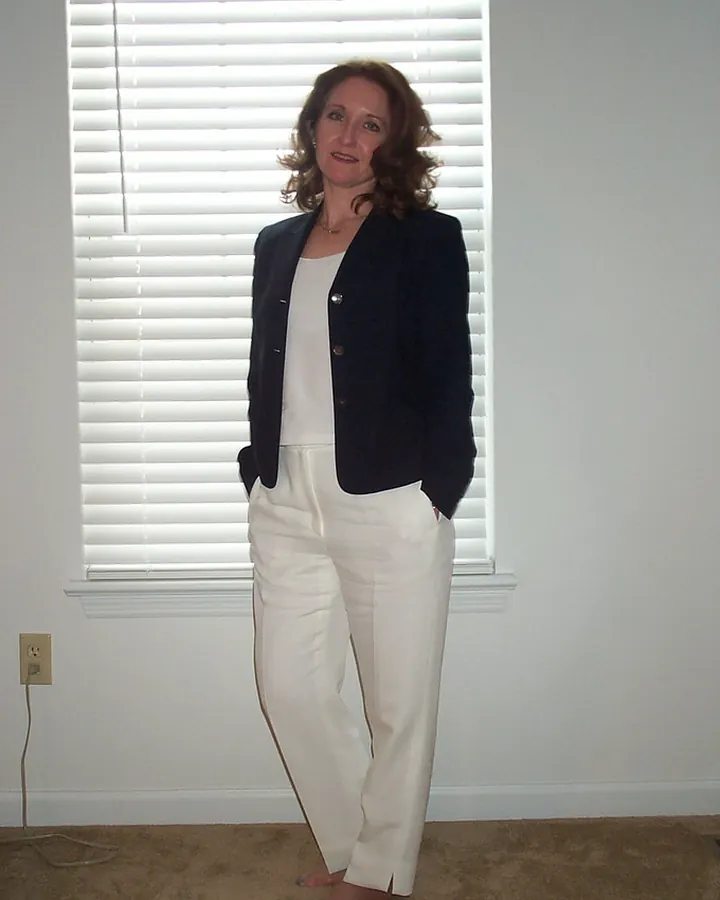 Femme aux cheveux bruns en costume noir et pantalon blanc, pieds nus, debout devant fenêtre à persiennes