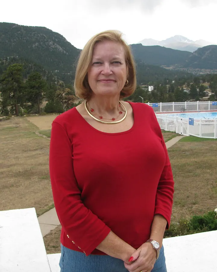 Femme aux cheveux blonds courts, en pull rouge, souriant à l'appareil, avec une piscine et des montagnes en arrière-plan