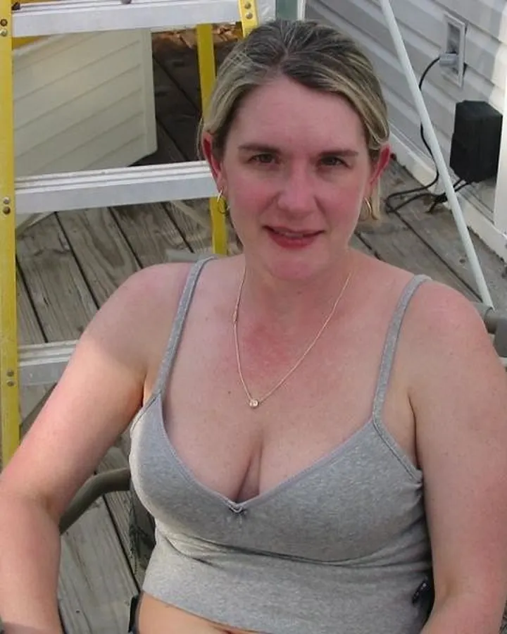 Femme aux cheveux blonds courts, en haut gris, souriant à l'appareil, assise sur une terrasse en bois