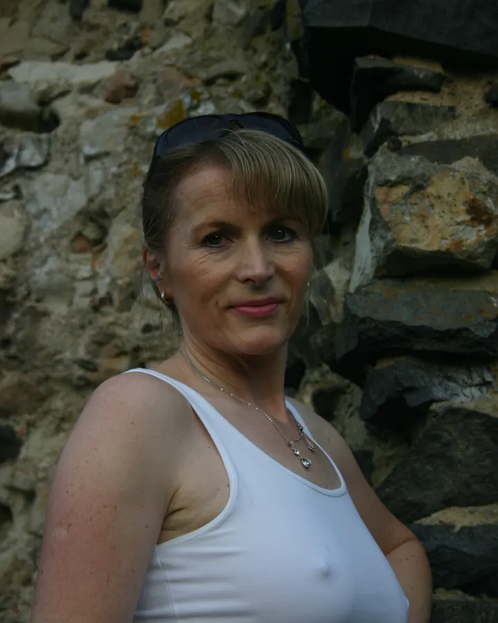 Femme aux cheveux blonds portant un haut blanc, debout contre un mur de pierre avec des lunettes de soleil sur la tête.