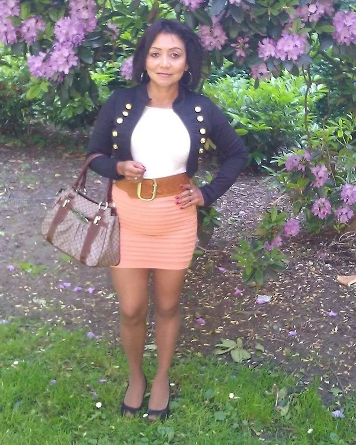 Femme aux cheveux noirs portant un haut noir et un short blanc, posant dans un jardin avec des fleurs roses.