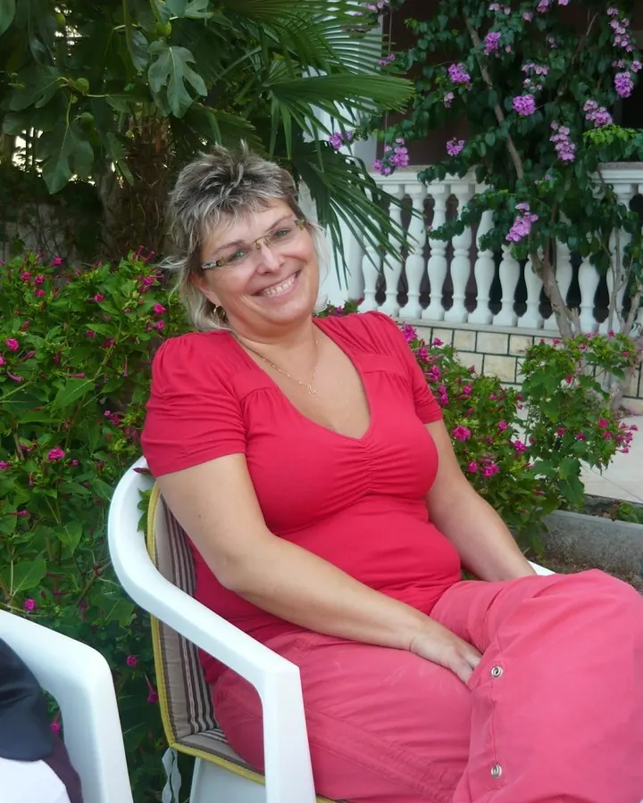 Femme aux cheveux gris, en robe rouge, assise sur une chaise blanche devant des fleurs roses