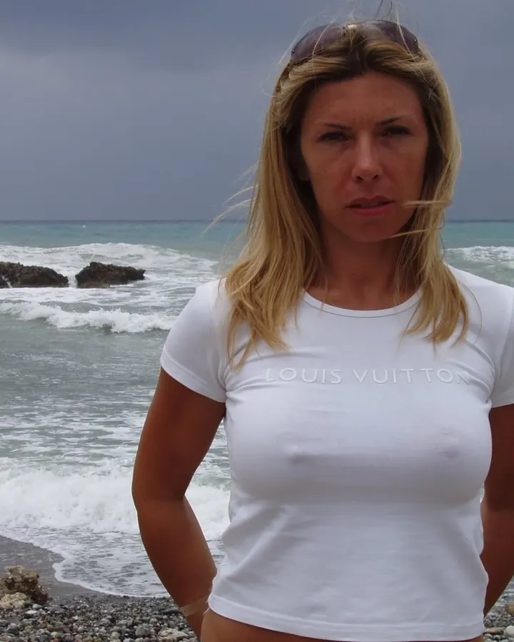 Femme blonde en t-shirt blanc Louis Vuitton sur une plage rocheuse, regard intense vers l'objectif.