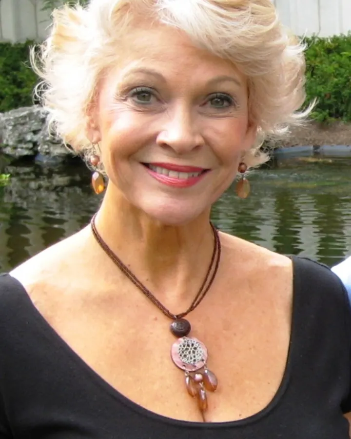 Femme aux cheveux blonds courts souriant, portant un haut noir et un collier avec un pendentif rond, devant un étang.